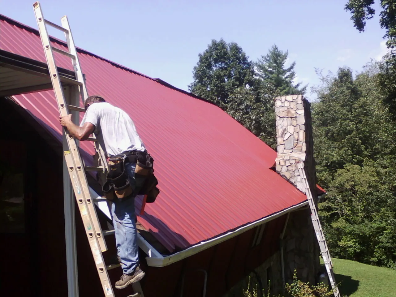 Roofer on a ladder beside a red metal roof and stone chimney — Soffit & Fascia Repair by Roofing Brothers in Malo